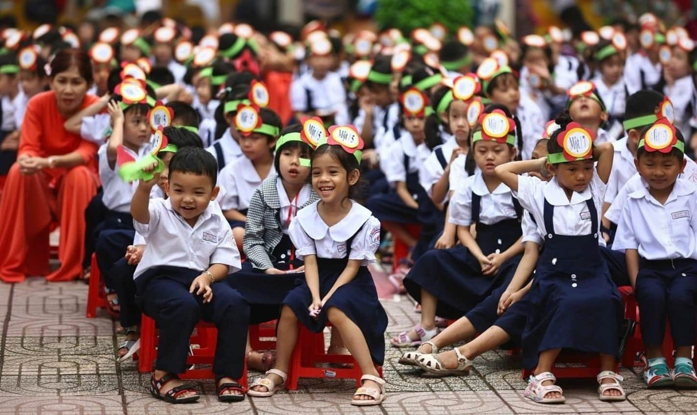 A group of Vietnamese schoolchildren in white-and-navy uniforms seated outdoors, wearing celebratory paper crowns. Two children in the foreground smile and look toward the camera.