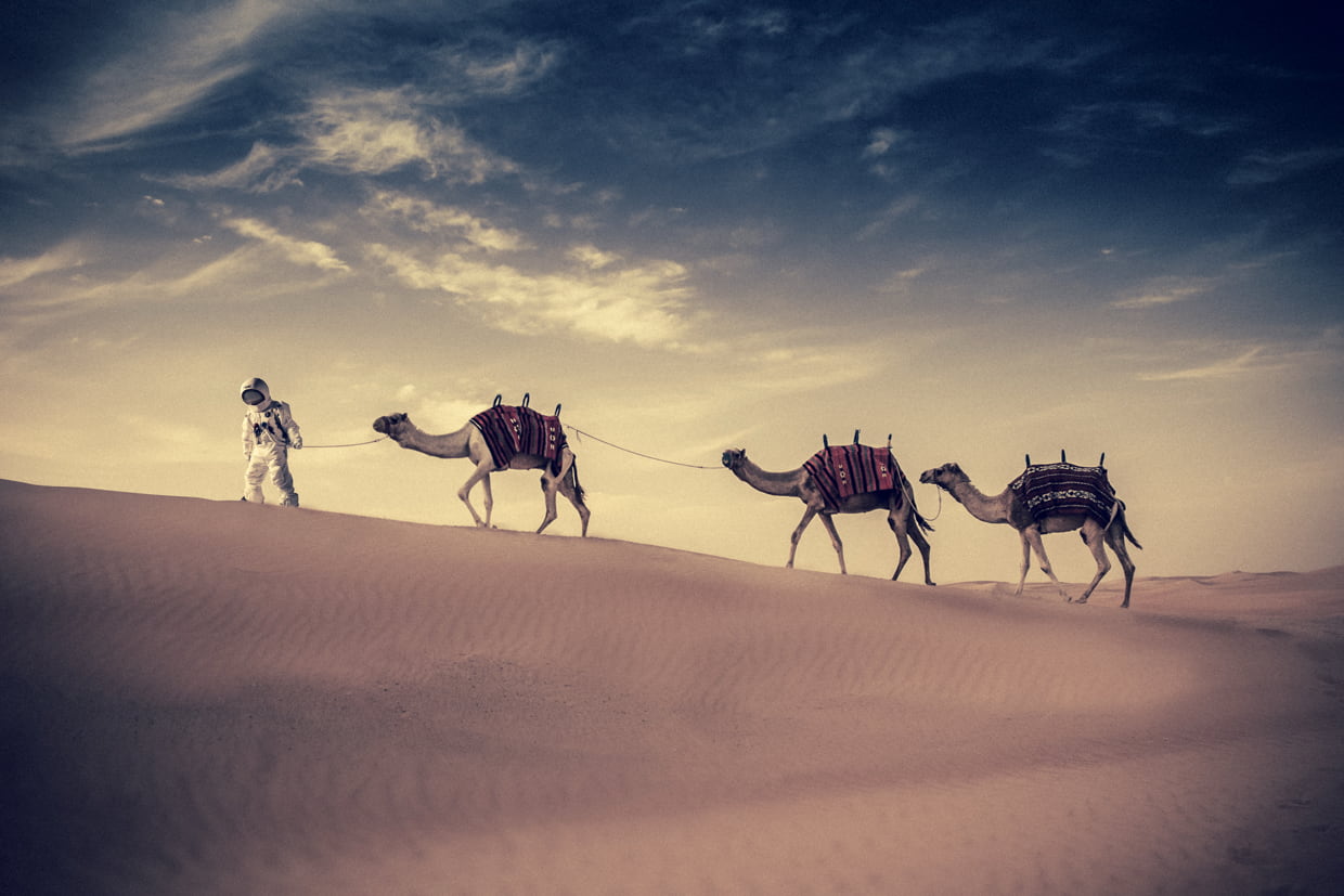 AXE astronaut walking across desert dunes with 3 camels.