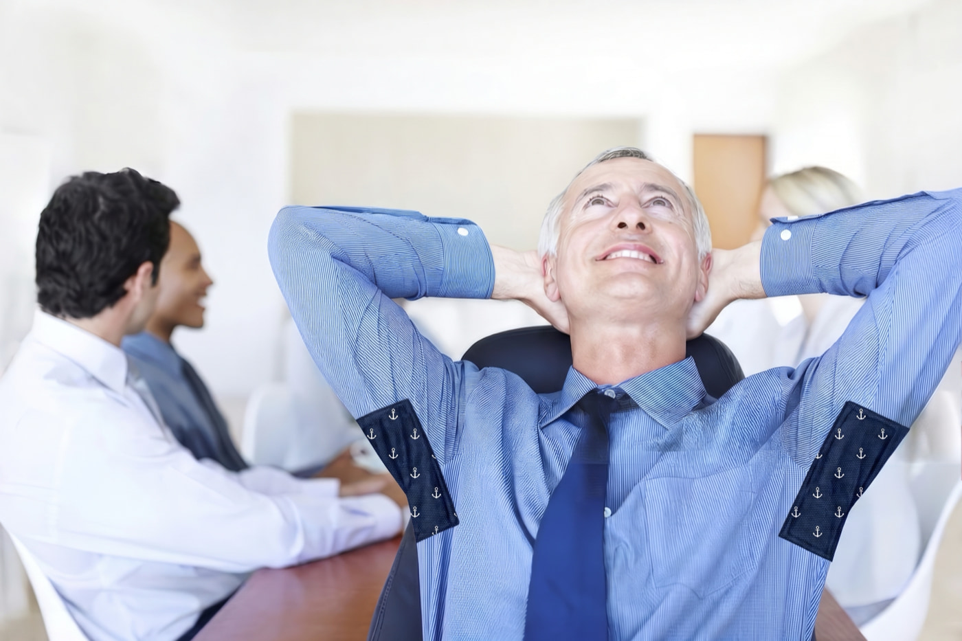 An executive leans back in a chair wearing visible underarm patches on both sleeves while colleagues sit blurred in the background.