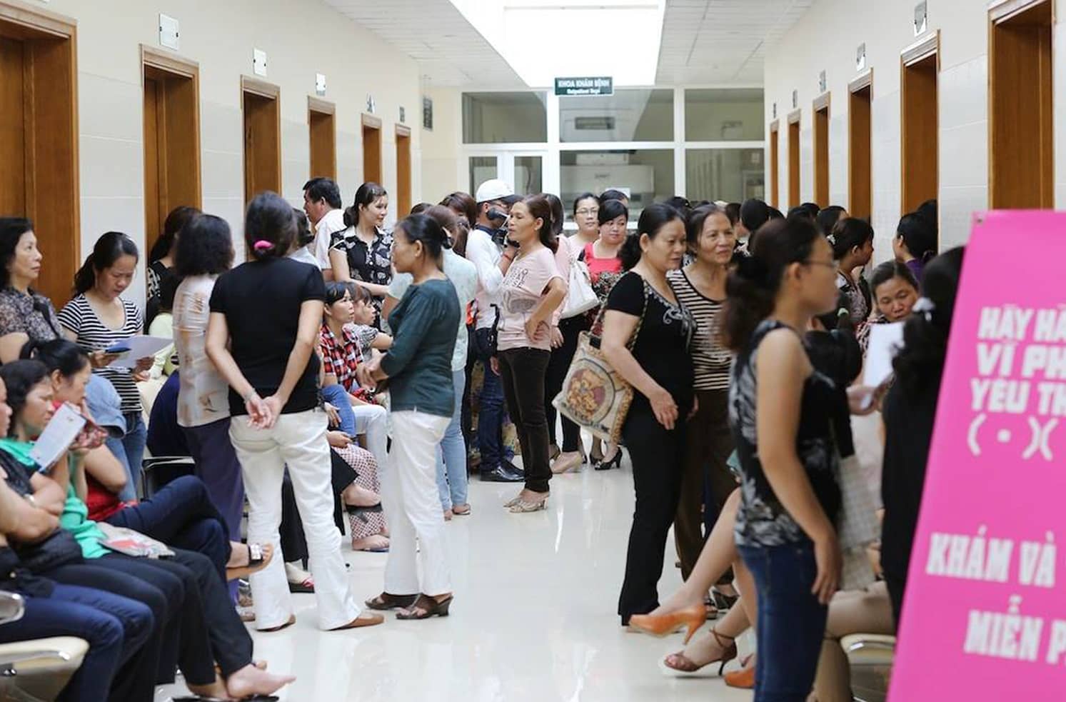 Crowded hospital hallway with women waiting near a pink We Care For Her screening sign.